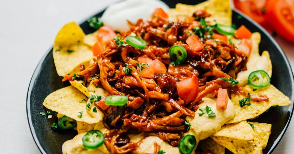 Vegan nachos on a black plate on a white background - These vegan nachos are piled high with easy mushroom BBQ "pulled pork" and a cashew-based queso cheese sauce that will knock your dairy-free socks off.