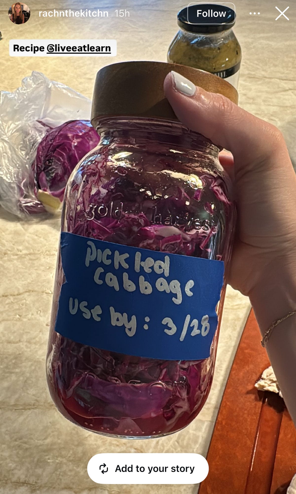 A hand holds a jar labeled pickled red cabbage, use by: 3/28. A halved red cabbage and a plastic bag rest on the counter.