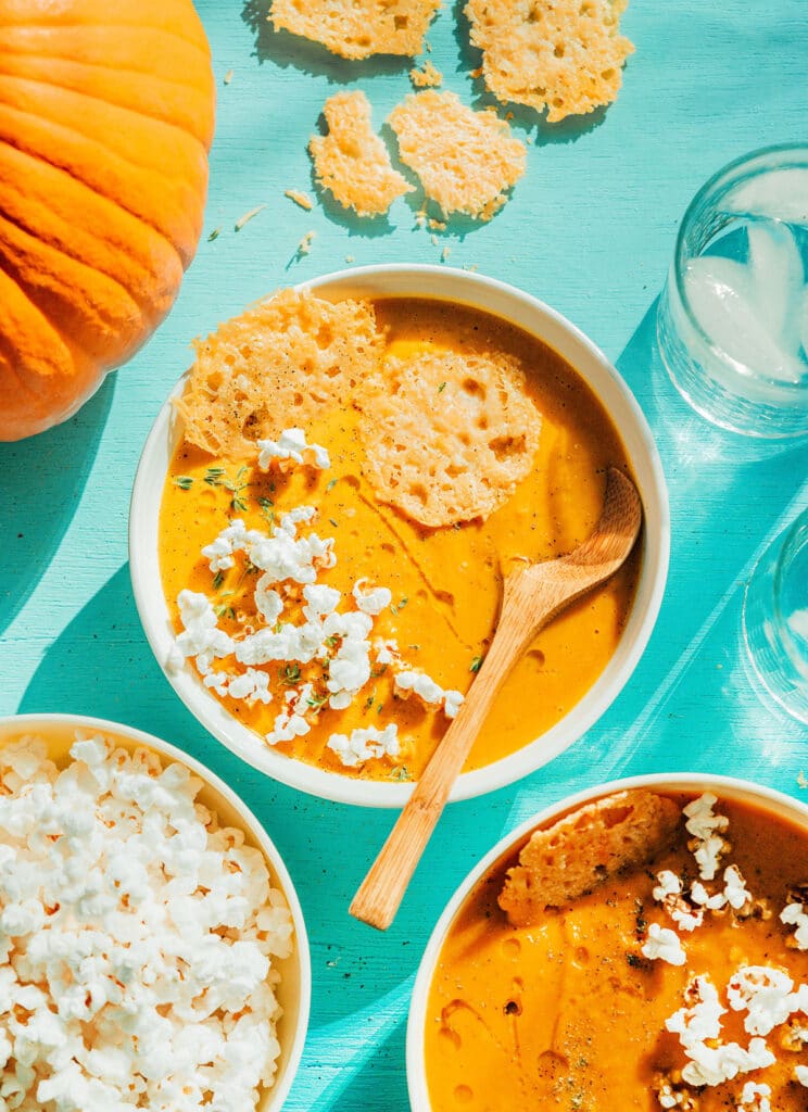 A white bowl filled with pumpkin soup and topped with parmesan crisps, popcorn, and spices
