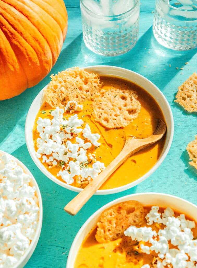 A white bowl filled with pumpkin soup and topped with parmesan crisps, popcorn, and spices