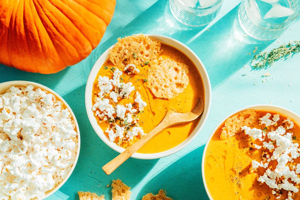 Two white bowls filled with pumpkin soup with parmesan popcorn crunch surrounded by ingredients