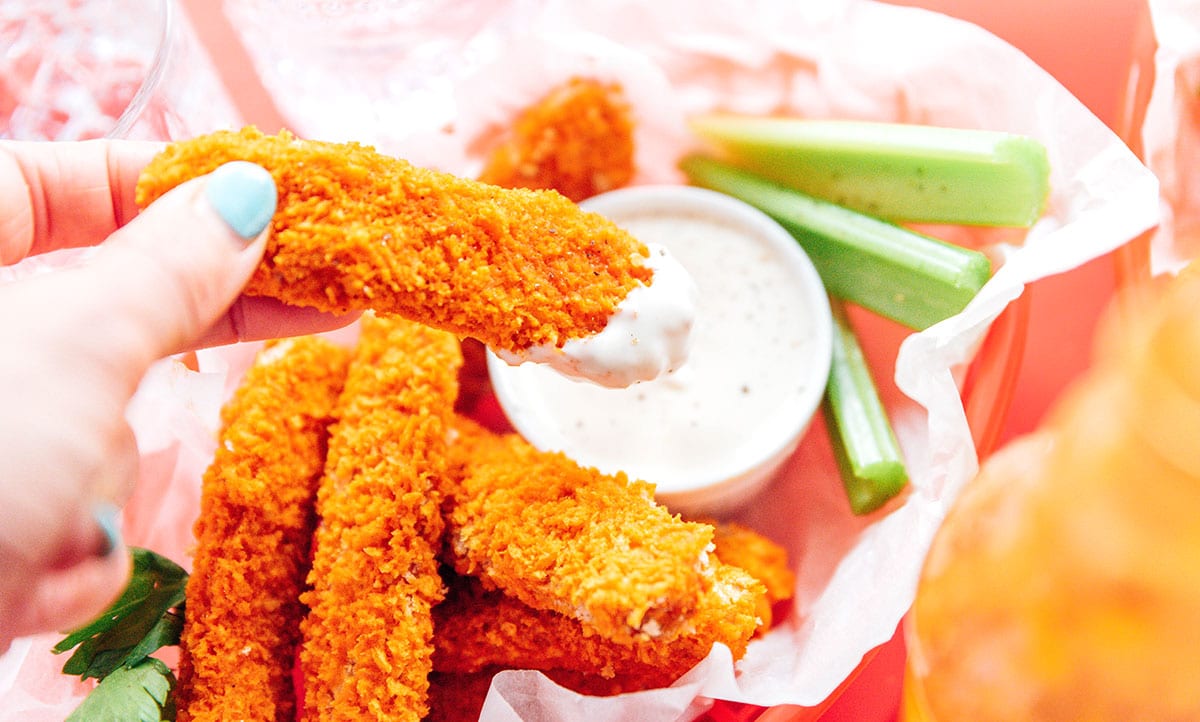 A hand dipping tempeh tenders in a red basket with ranch dip and celery sticks