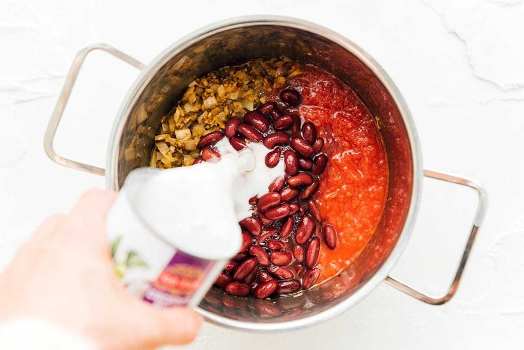 Pouring coconut milk into a sauce pan filled with tomato sauce, kidney beans, and crushed tomatoes