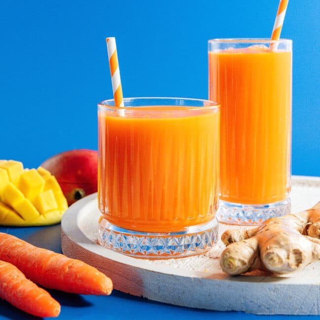 Carrot juice in two classes with ginger and mango nearby.
