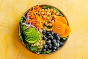 A bowl inspired by How To Build A Buddha Bowl: spinach, quinoa, avocado slices, shredded carrots, red onion, chickpeas, orange slices, and blueberries on a yellow background.