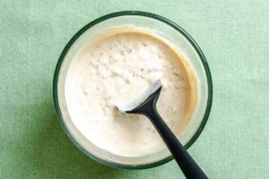 A glass bowl of creamy French onion dip with visible herbs or seasonings, being stirred with a black spatula, on a green textured surface.