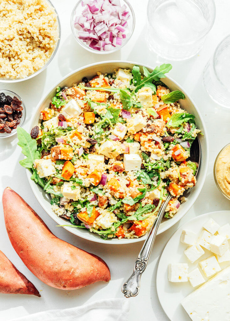 A white bowl filled with sweet potato salad with quinoa and hummus dressing surrounded by various bowls of ingredients