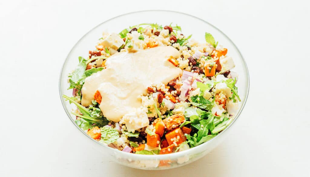 A clear glass bowl filled with sweet potato salad topped with hummus dressing