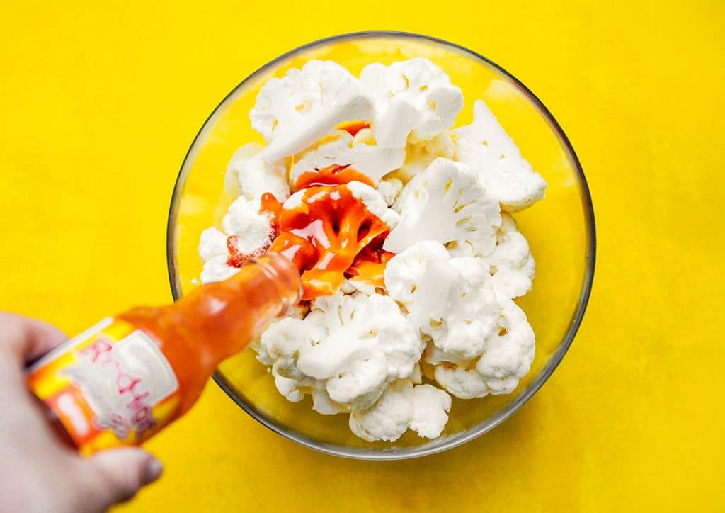 Pouring buffalo sauce into a bowl of cauliflower on a yellow background