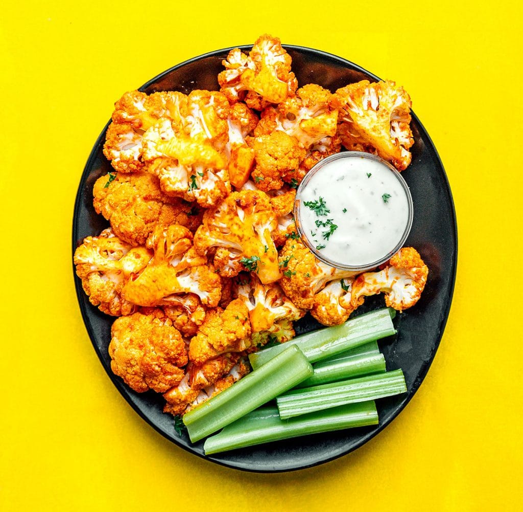 Buffalo cauliflower, celery, and dip on a black plate on a yellow background