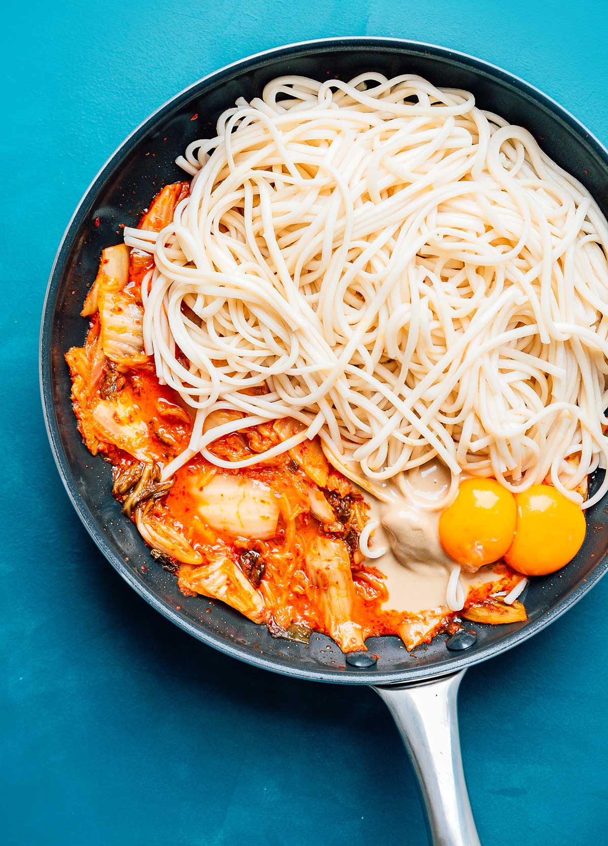 Kimchi topped with udon noodles and egg yolks in a sauce pan on a blue background