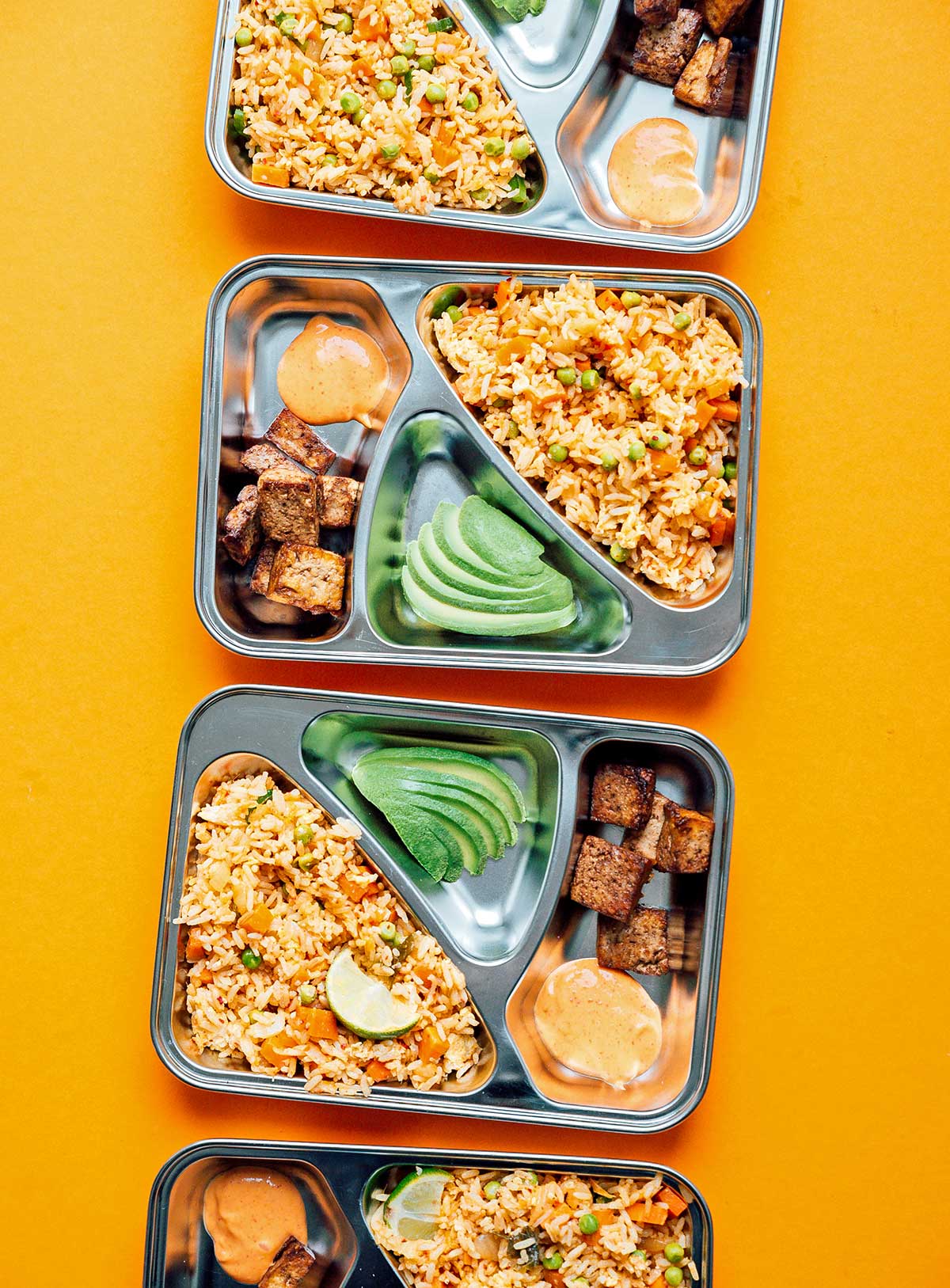 Four meal prep containers filled with kimchi fried rice, tofu, and avocado slices on a yellow background