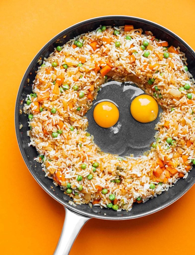 Vegetables, kimchi, rice, and eggs cooking in a skillet on an orange background