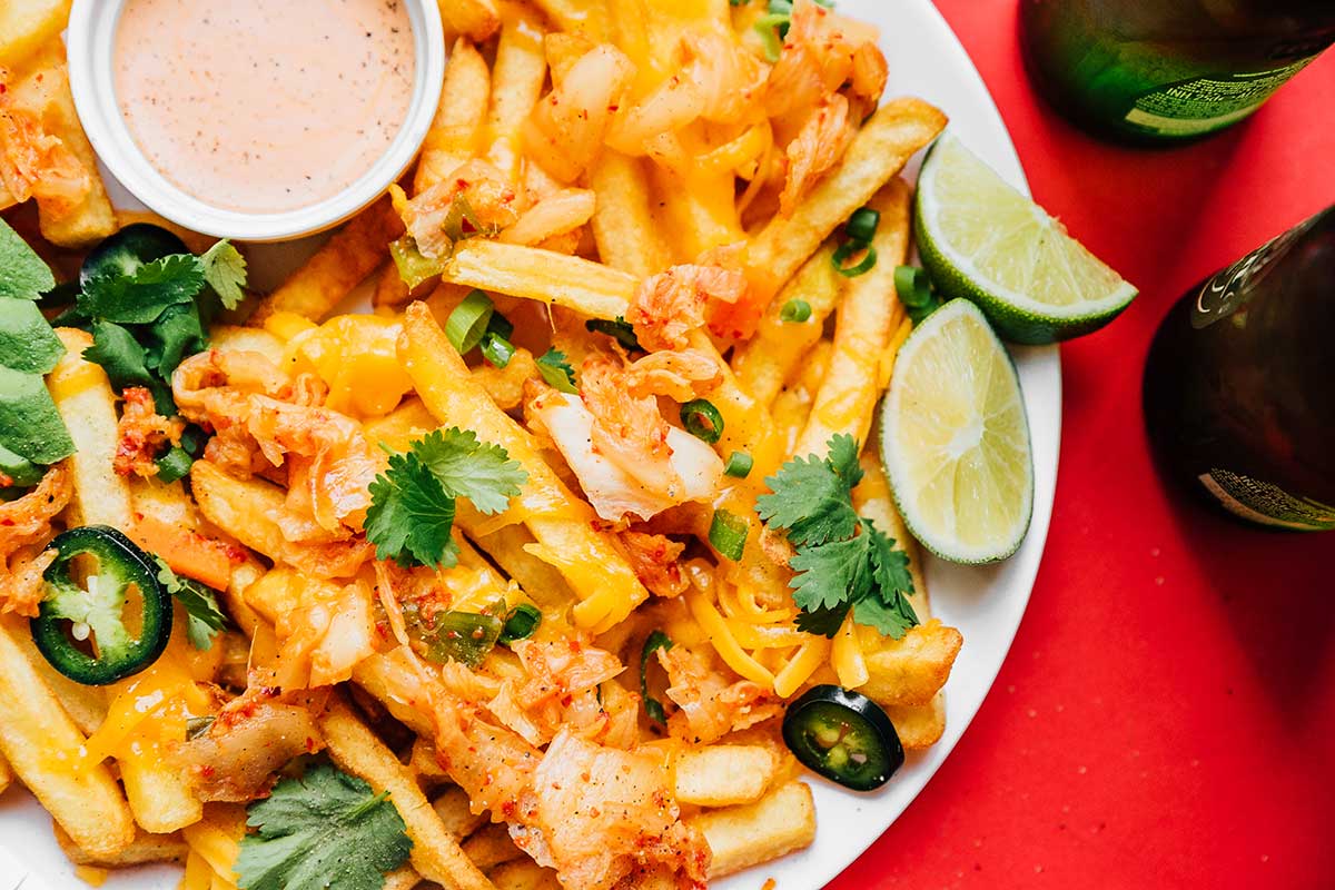 Close up shot of kimchi fries, sauce, lime slices, and toppings on a plate on a red background