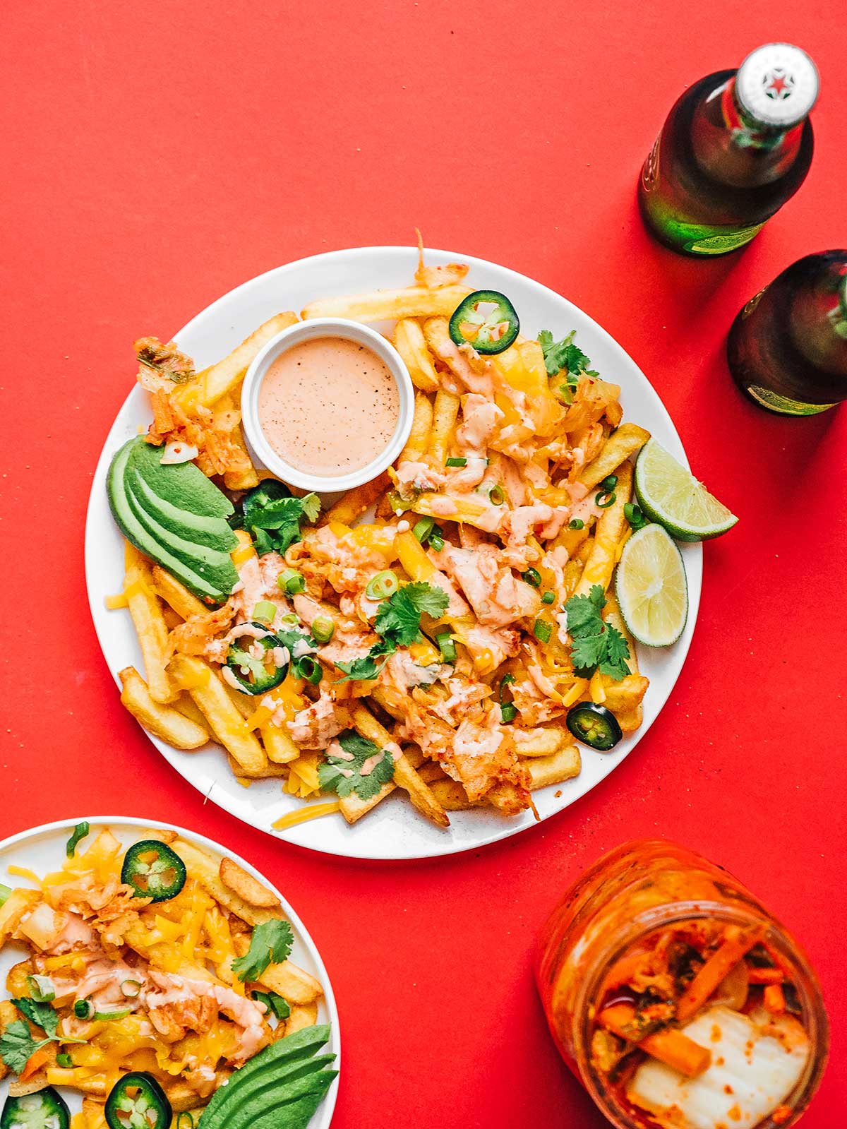 Bird's eye photo of kimchi fries, sauce, avocado slices, lime slices, and toppings on a plate on a red background