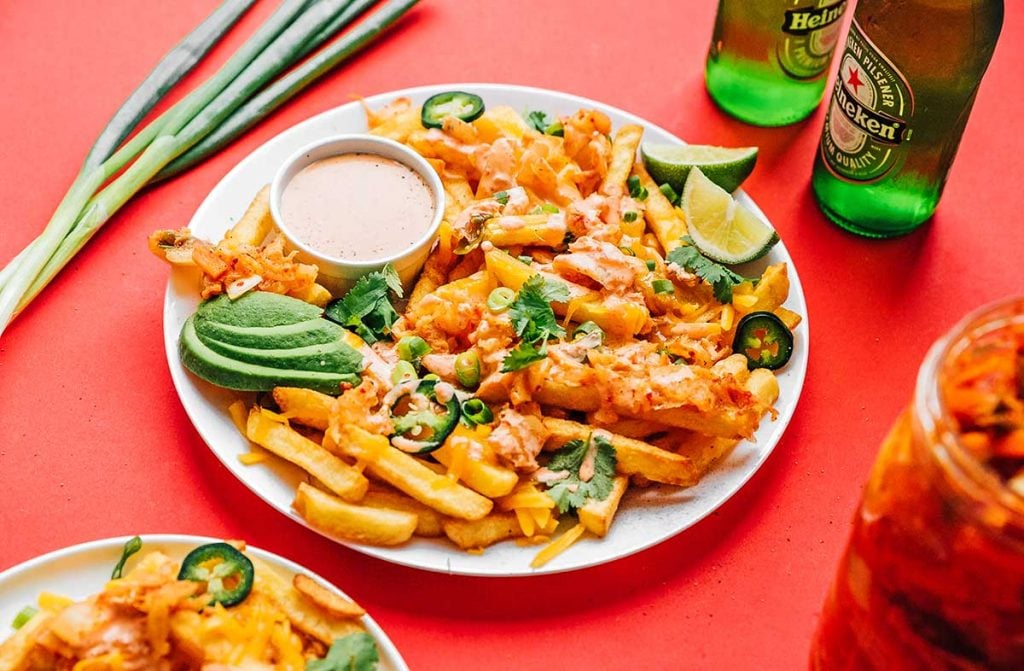Kimchi fries, sauce, avocado slices, lime slices, and toppings on a plate on a red background