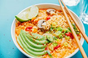 A bowl of kimchi ramen with sliced avocado, mushrooms, lime wedges, green onions, and red chili flakes on a blue surface, accompanied by wooden chopsticks.