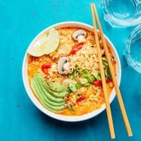 Bowl of kimchi ramen with avocado slices, mushrooms, lime wedge, and green onions on a blue surface. Chopsticks and two glasses of water are nearby.