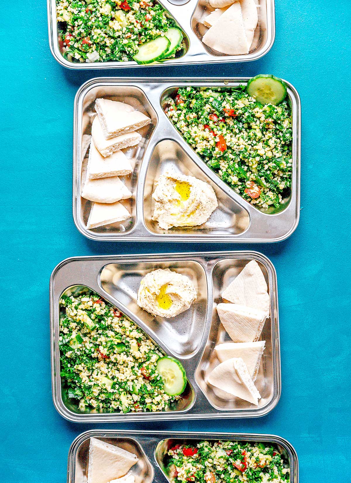 Four meal prep containers on a blue background filled with tabbouleh, hummus, and pita bread