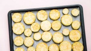 A baking tray with evenly arranged round slices of roasted vegetables, possibly zucchini or squash, placed on parchment paper. Perfectly roasted to complement a creamy side like baba ganoush.
