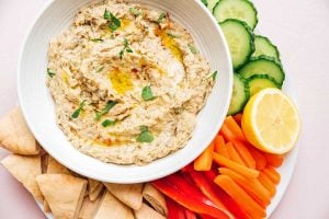 A bowl of hummus garnished with herbs and olive oil, accompanied by baba ganoush, is surrounded by sliced cucumber, lemon, carrot sticks, red bell peppers, and pita bread pieces on a plate.