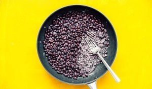 A frying pan on a yellow surface contains black beans, partially mashed with a fork, perfect for a hearty huevos rancheros breakfast.