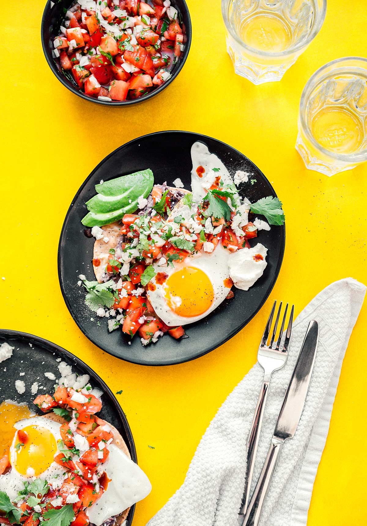 Two plates of huevos rancheros accompanied by two glasses of water and a bowl of pico de gallo