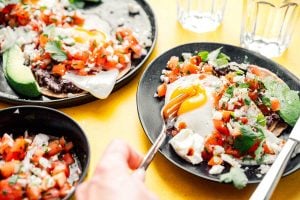 A vibrant yellow table hosts plates of huevos rancheros, brimming with eggs, salsa, beans, avocado, and cilantro. Glasses of water await in the background as a hand eagerly grips a fork near one plate.