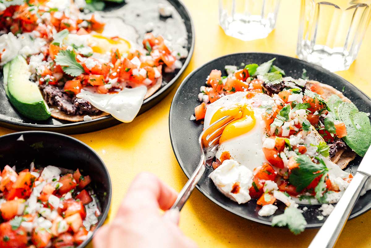 Two plates of huevos rancheros and a bowl of pico de gallo on a yellow background
