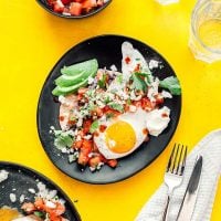 Two plates of sunny-side-up eggs styled as huevos rancheros with salsa, avocado, and cheese rest on a yellow tablecloth. A small bowl of salsa complements the scene, alongside two glasses of water, cutlery, and a napkin.