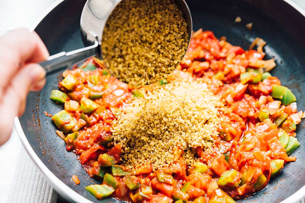 Adding bulgur to a pan filled with chopped veggies and diced tomatoes.
