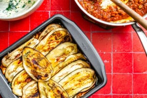 A baking dish filled with layered grilled eggplant slices sits on a red tiled surface next to a pan of tomato sauce and a bowl of creamy mixture, ready to become a delicious homemade eggplant lasagna.