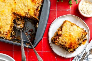 A baked eggplant lasagna with a cheesy top in a pan, a serving spoon, and a square piece plated on a white plate next to a small bowl of shredded cheese on a red tiled surface.