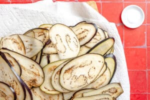 Thin slices of raw eggplant, perfect for eggplant lasagna, are arranged on a white towel, with a small dish of salt on a red tiled surface nearby.