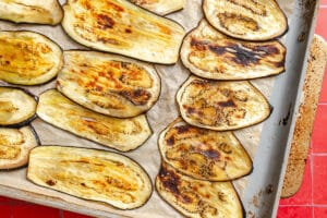 Slices of roasted eggplant, perfect for eggplant lasagna, are arranged on a parchment-lined baking sheet, showing some charred and golden-brown areas.