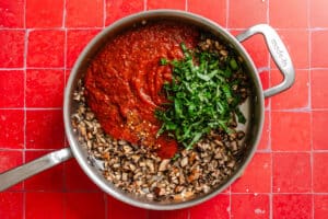A pan on a red tiled surface contains chopped mushrooms, tomato sauce, and freshly chopped basil, ready to be mixed for a delicious eggplant lasagna.