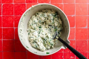 A mixing bowl filled with thick, creamy batter for eggplant lasagna, packed with chopped greens and herbs, sits on a red tiled countertop. A black spoon rests in the bowl.