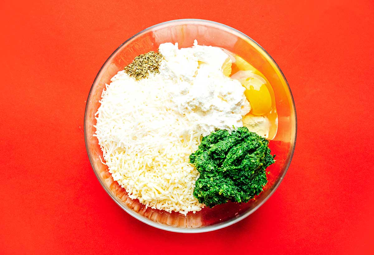 Various grated cheeses, ricotta, mozzarella, egg, spinach, and spices in a clear bowl ready to be mixed