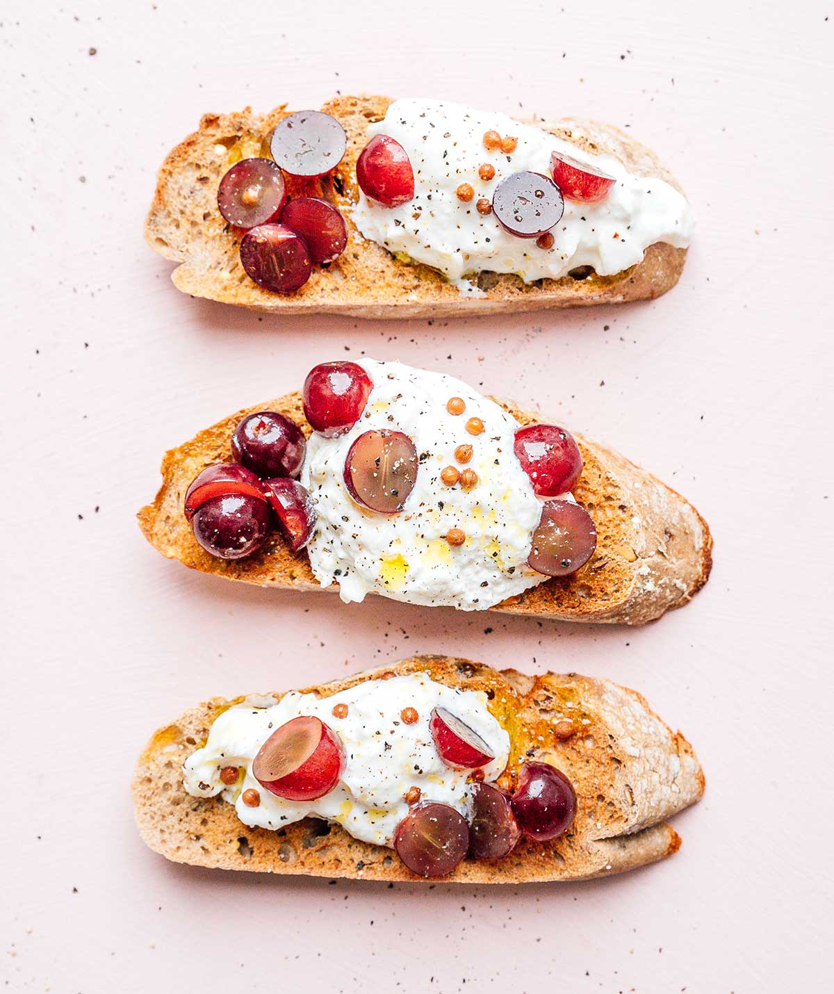 Three slices of toast topped with burrata, black peppercorns, and pickled grapes