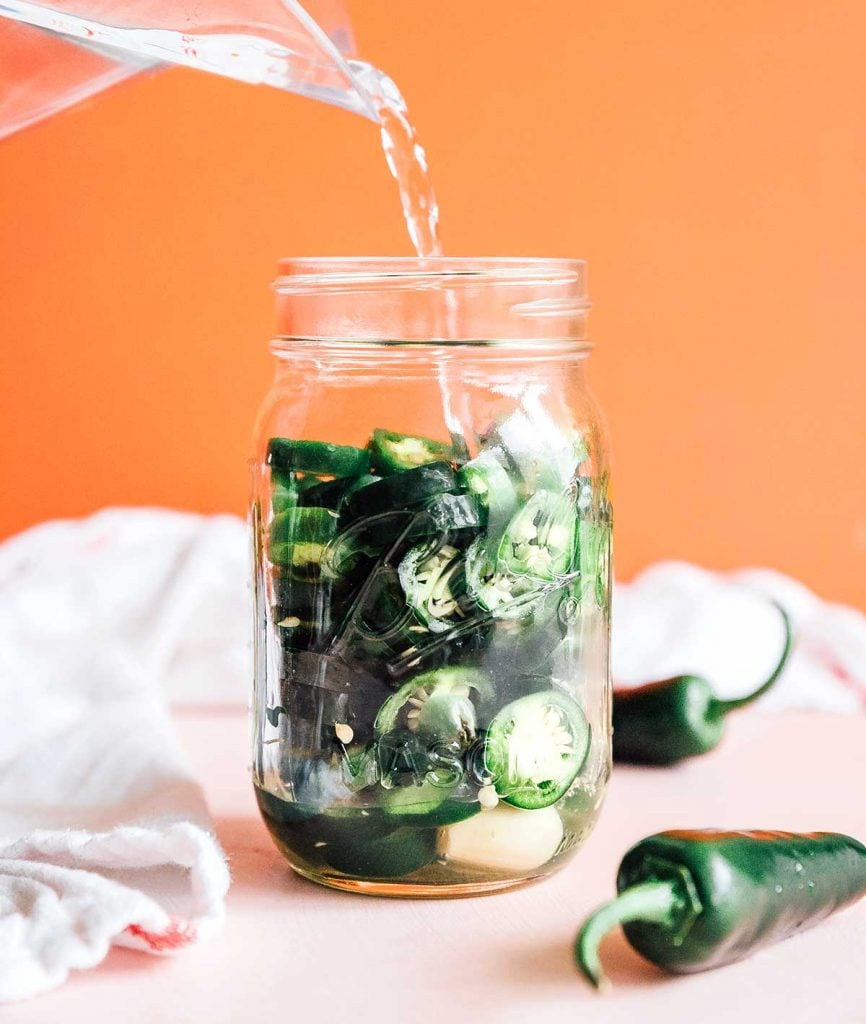 Pouring the vinegar pickling mixture into a mason jar filled with sliced jalapeños