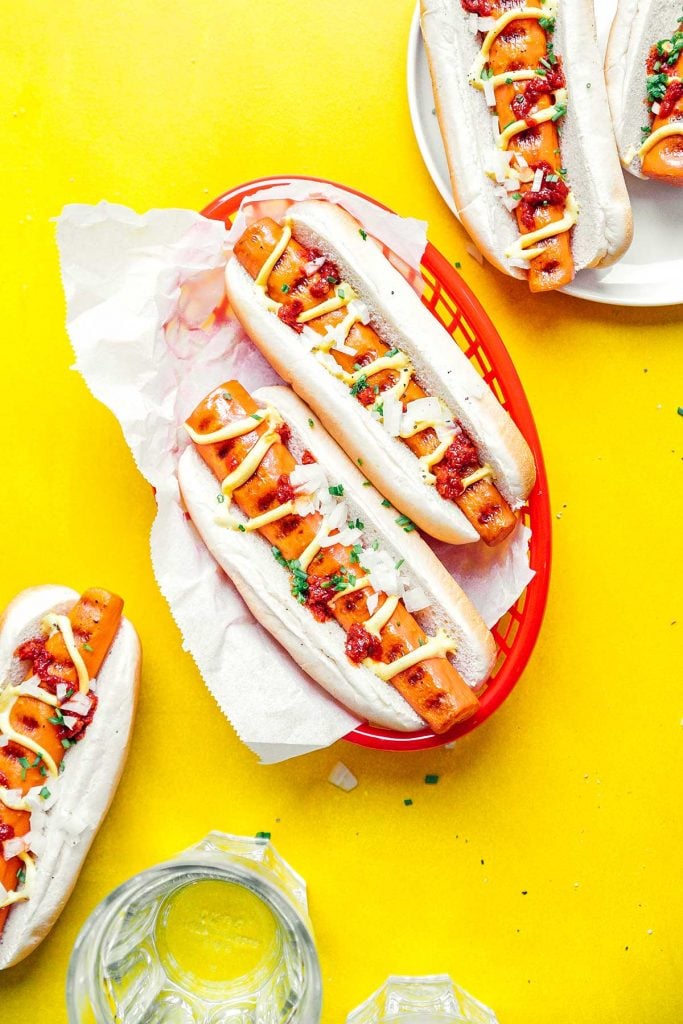 A basket filled with two vegetarian carrot hot dogs on a yellow background