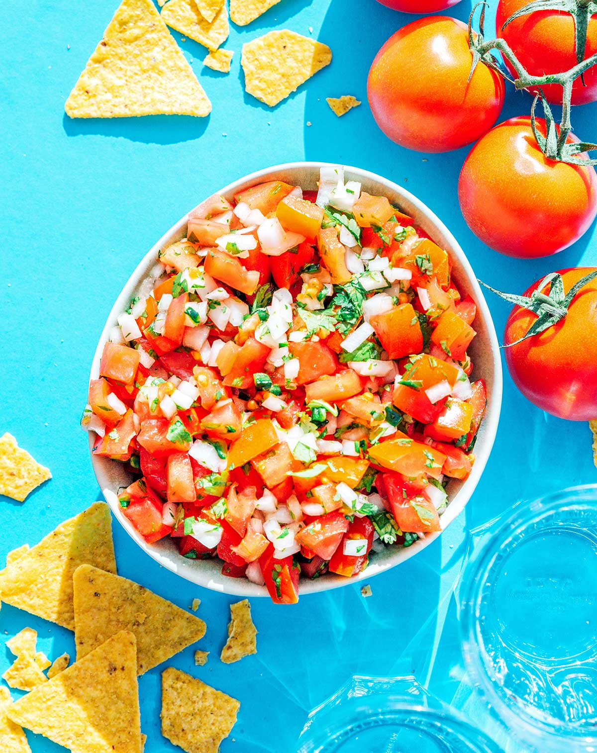 Pico de gallo in a white bowl with chips and tomatoes scattered around