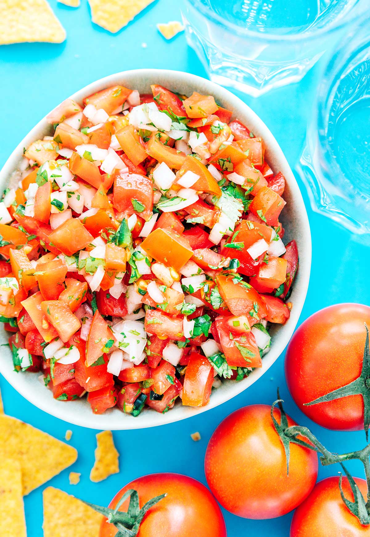 Pico de gallo in a white bowl with chips and tomatoes scattered around