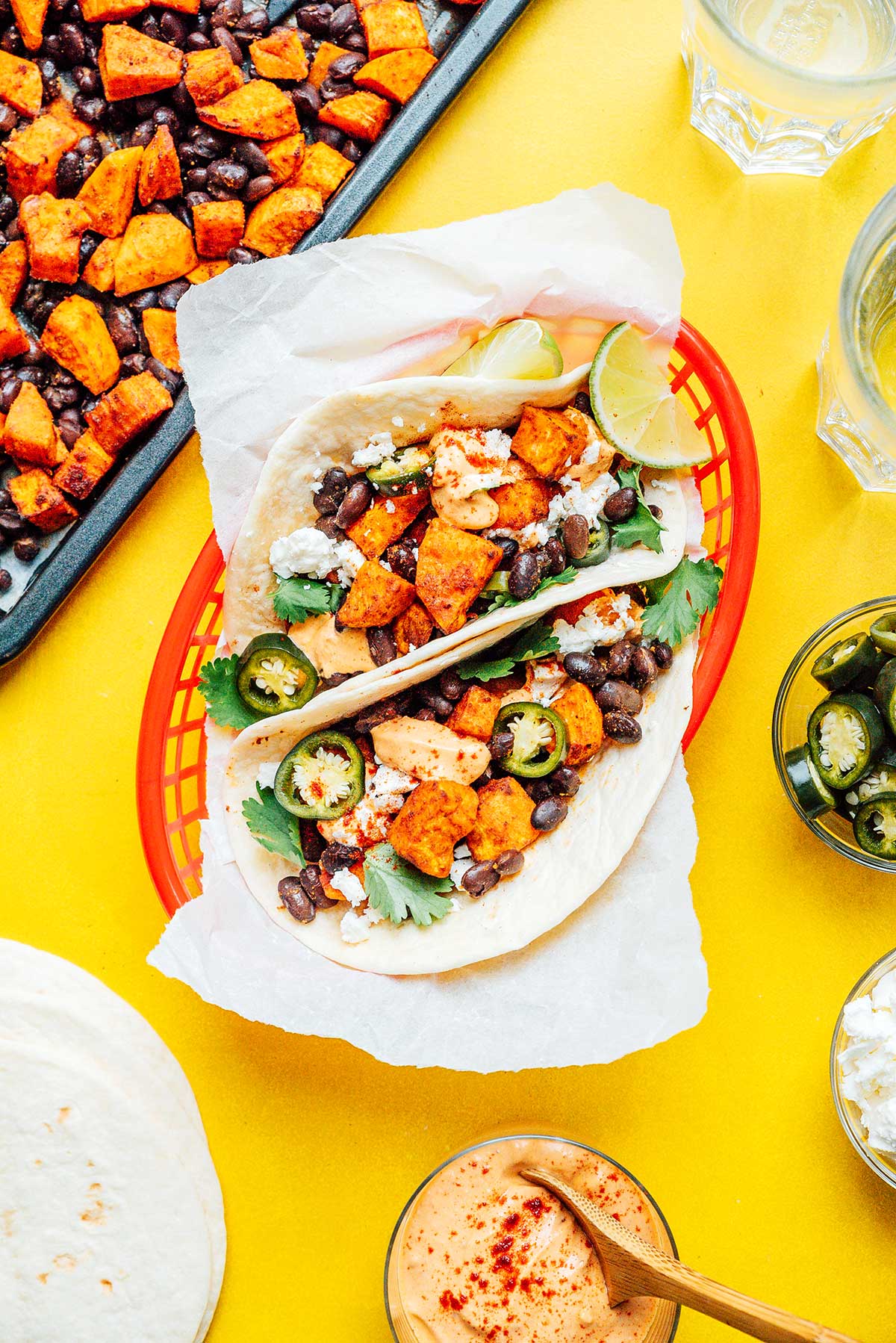 Two sweet potato tacos in a red basket surrounded by various taco ingredients 