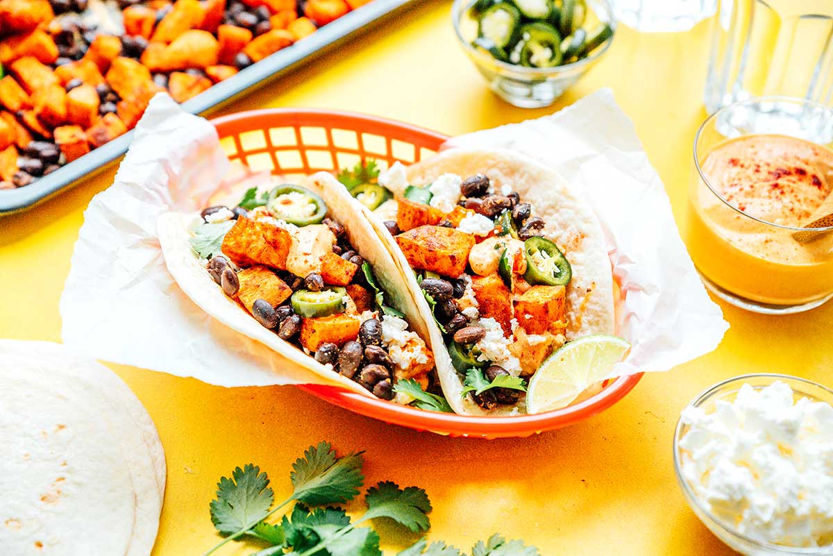 Two sweet potato tacos in a red basket surrounded by various taco ingredients 