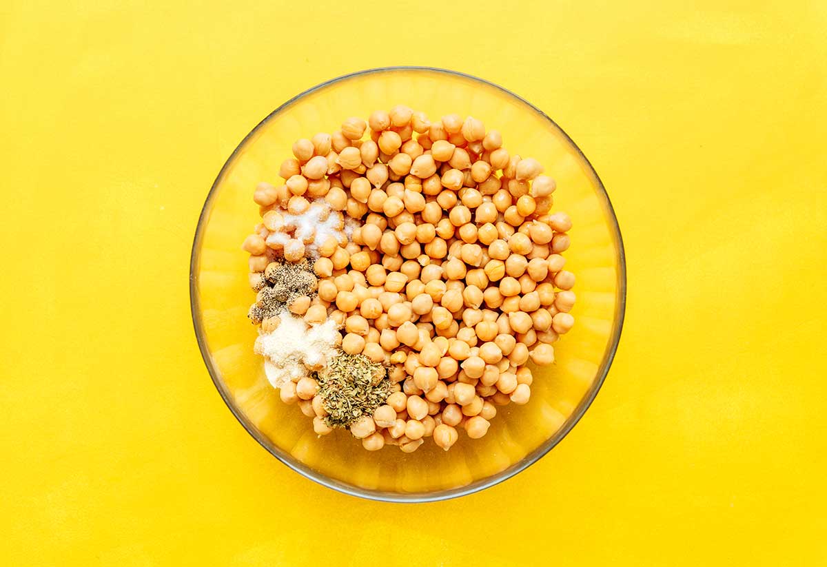 A clear glass bowl filled with chickpeas, oregano, garlic powder, salt, and pepper