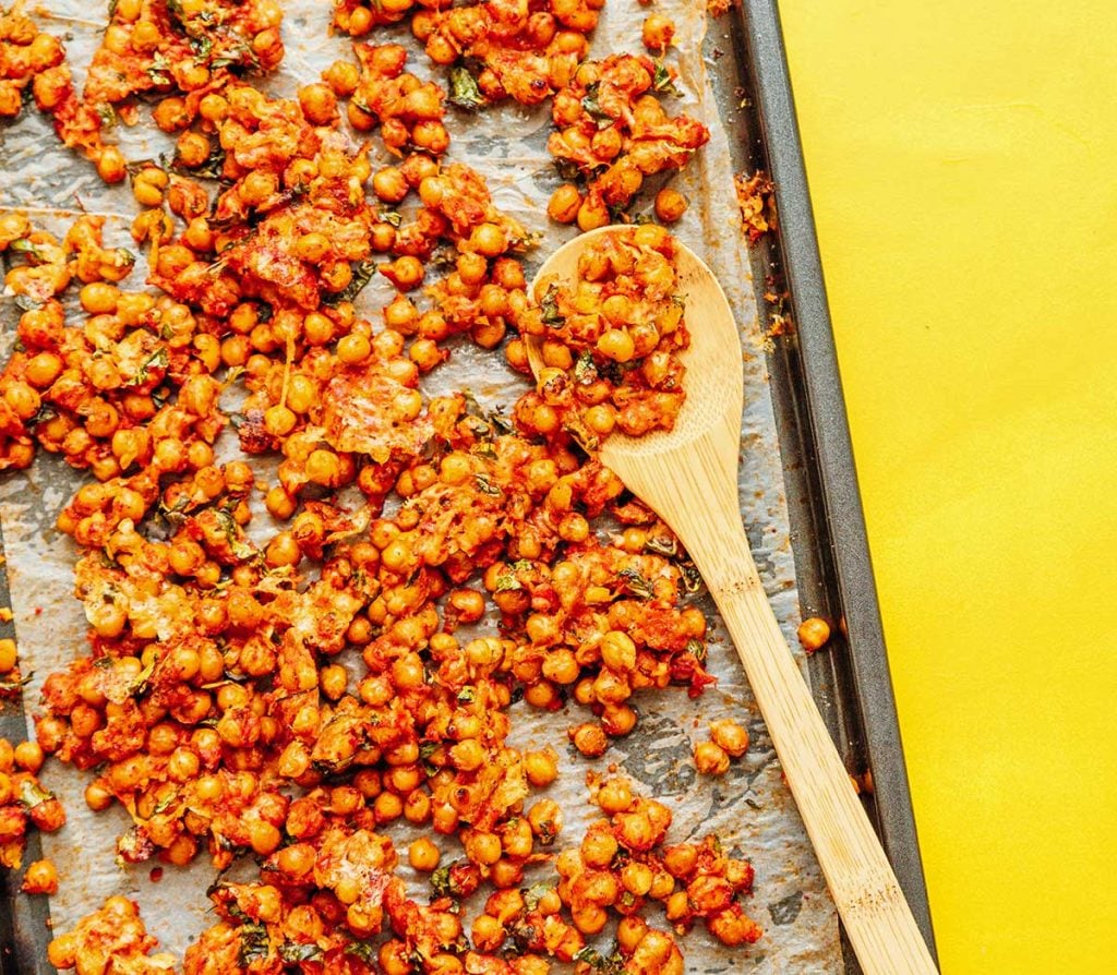 A wooden spoon scooping up a spoonful of chickpeas from a sheet pan filled with pizza roasted chickpeas 