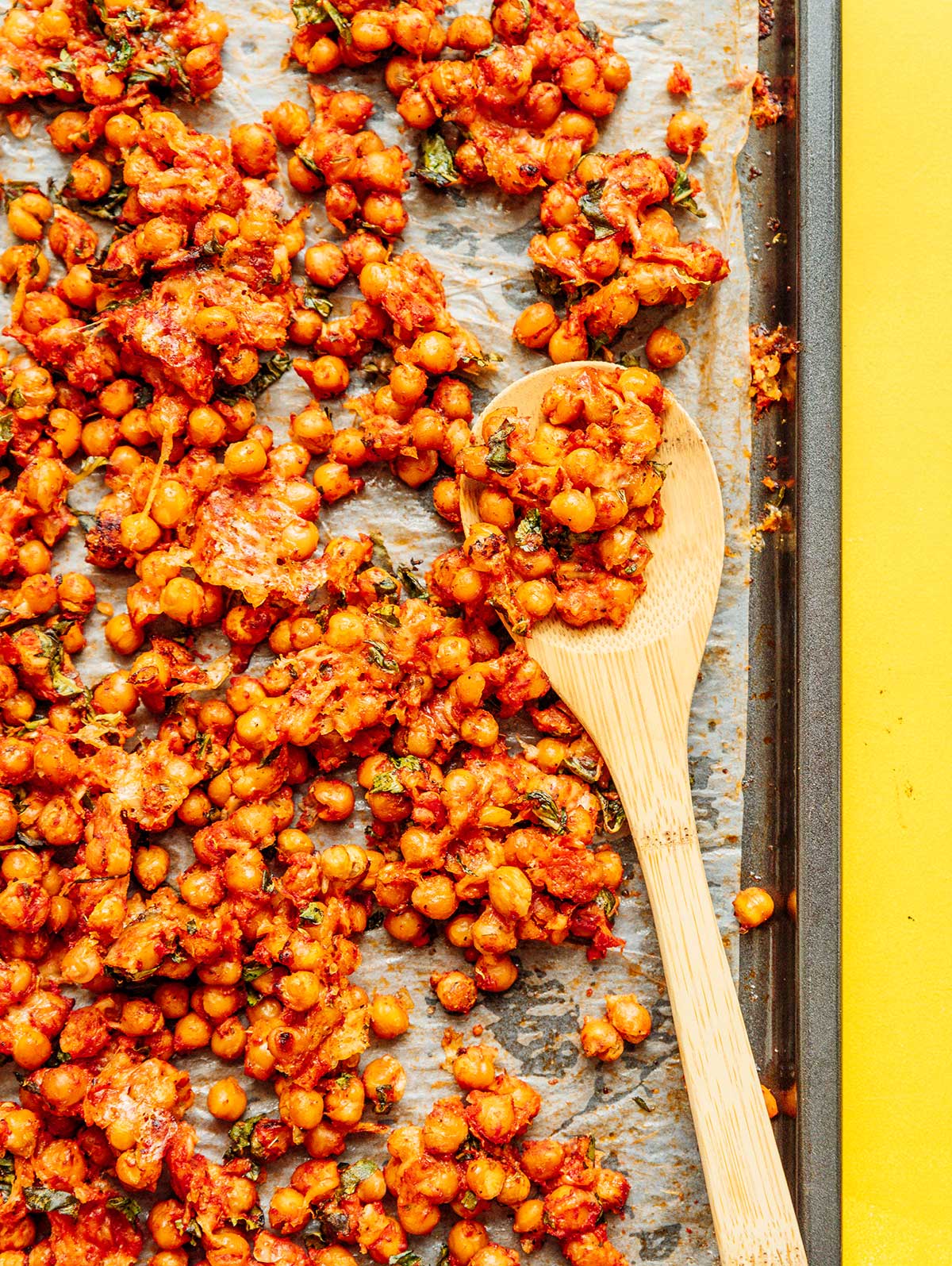 A wooden spoon scooping up a spoonful of chickpeas from a sheet pan filled with pizza roasted chickpeas 