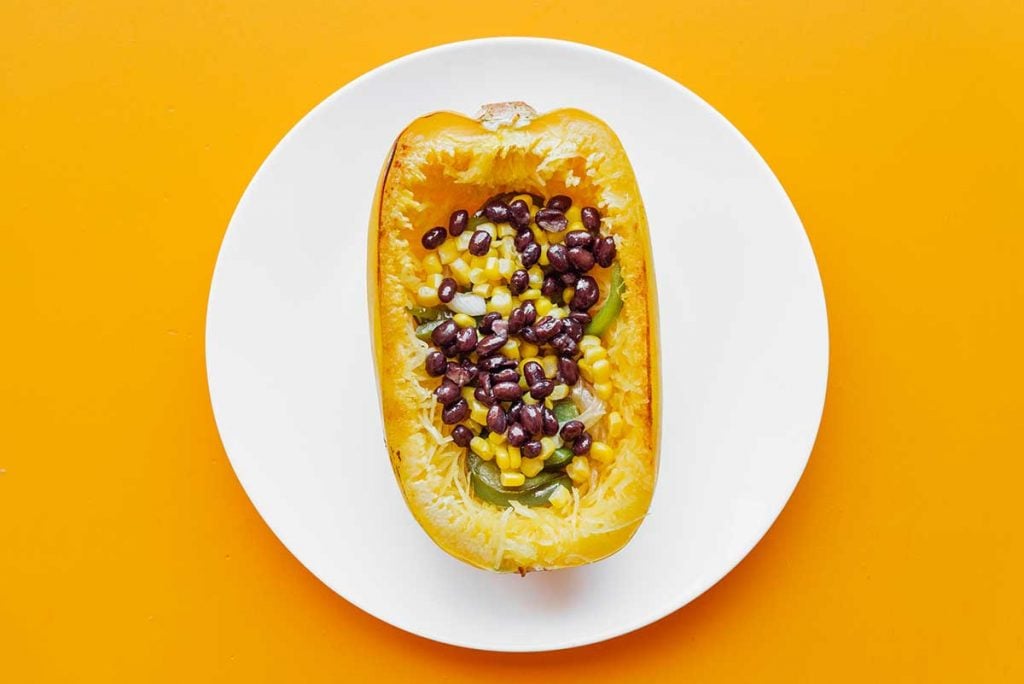 A hollowed out spaghetti squash half filled with sautéed green peppers and onions, corn, and black beans