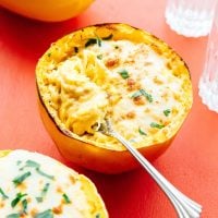 Spaghetti squash mac and cheese filled with creamy indulgence, garnished with chopped herbs, rests invitingly on a red surface. A fork sits ready to dive into the cheesy goodness.